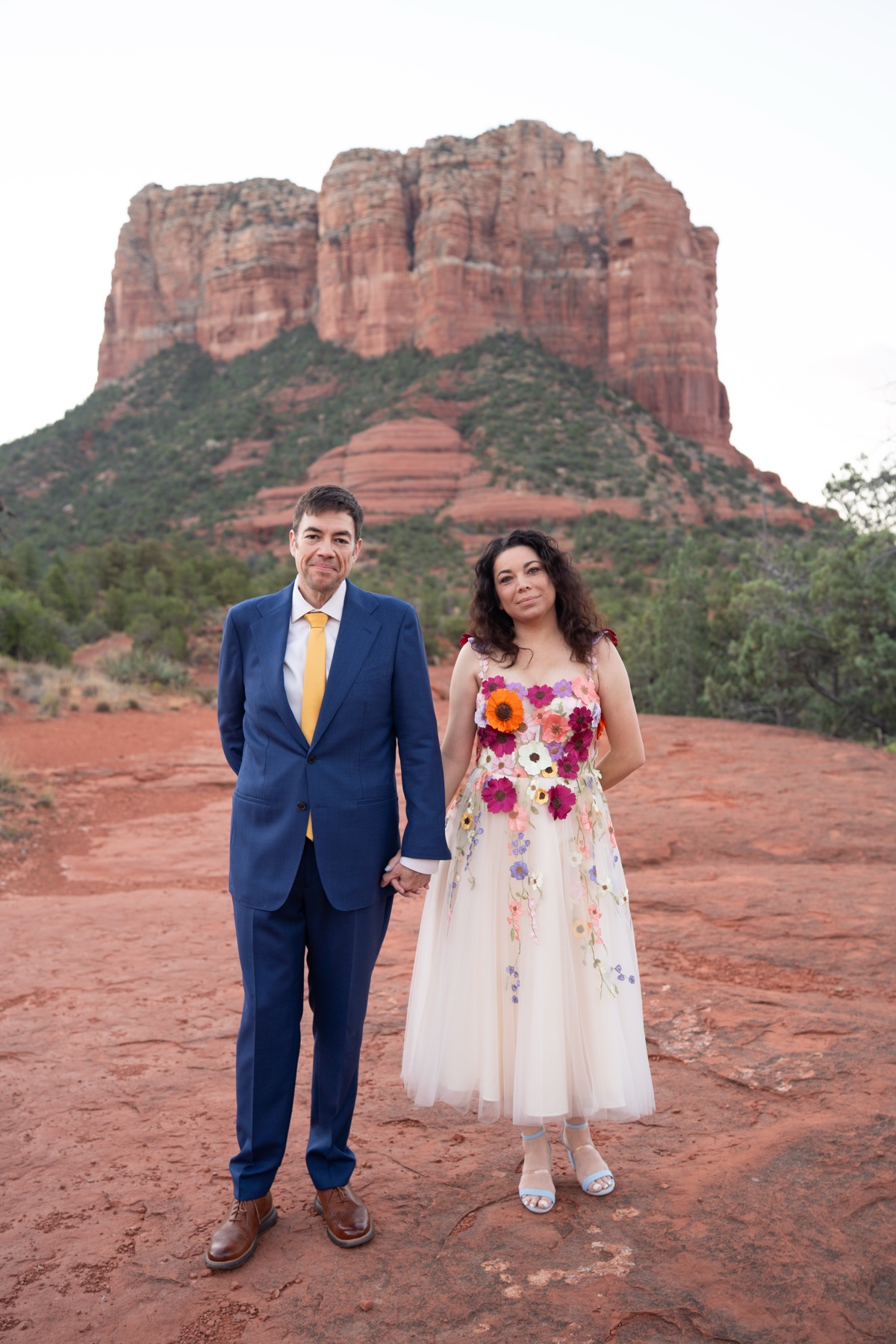 A Sedona elopement couple in colorful wedding attire standing beneath a red-rock butte