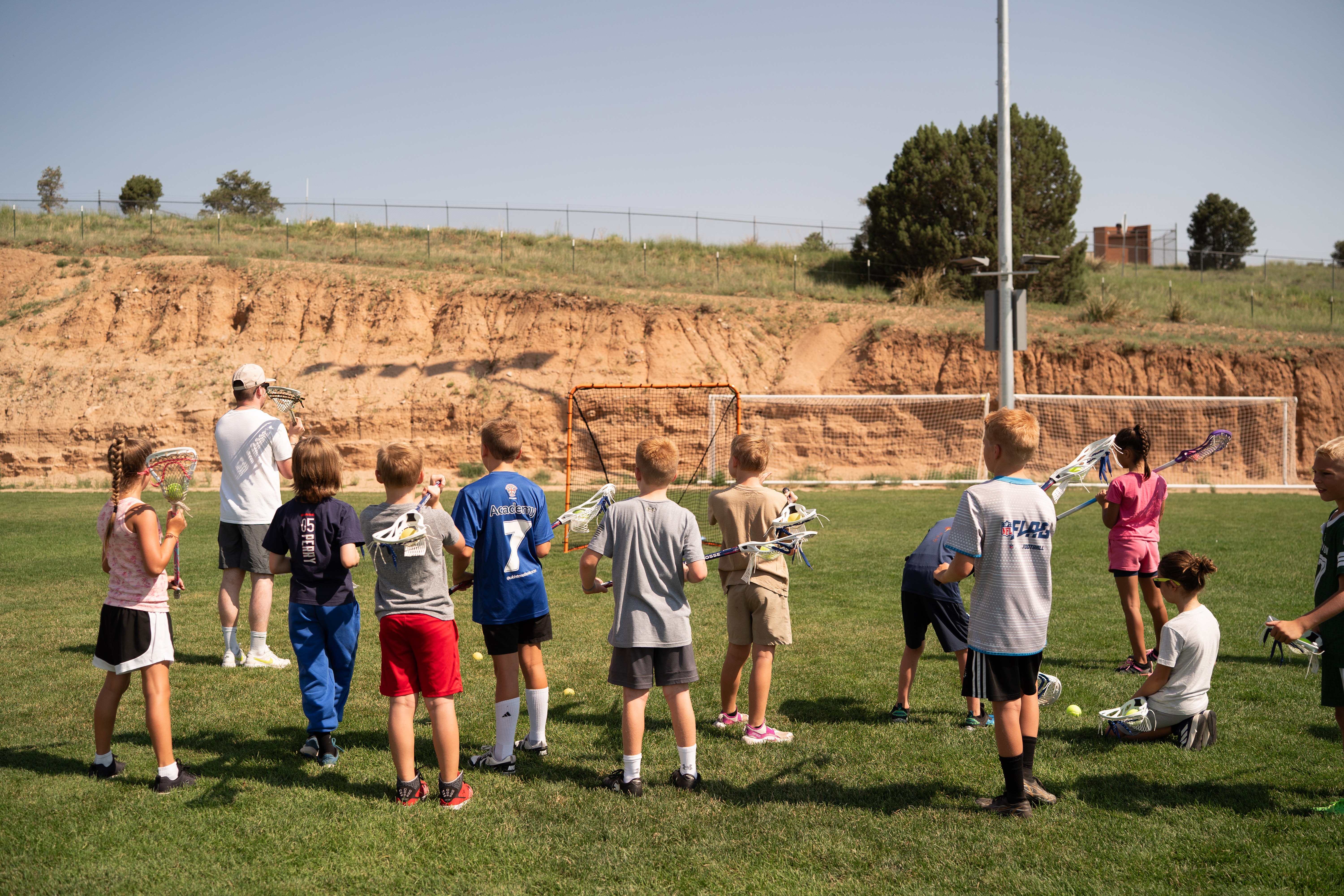 children gather around a coach on a green grassy field with lacrosse sticks in their hands intently listening to what the coach has to say