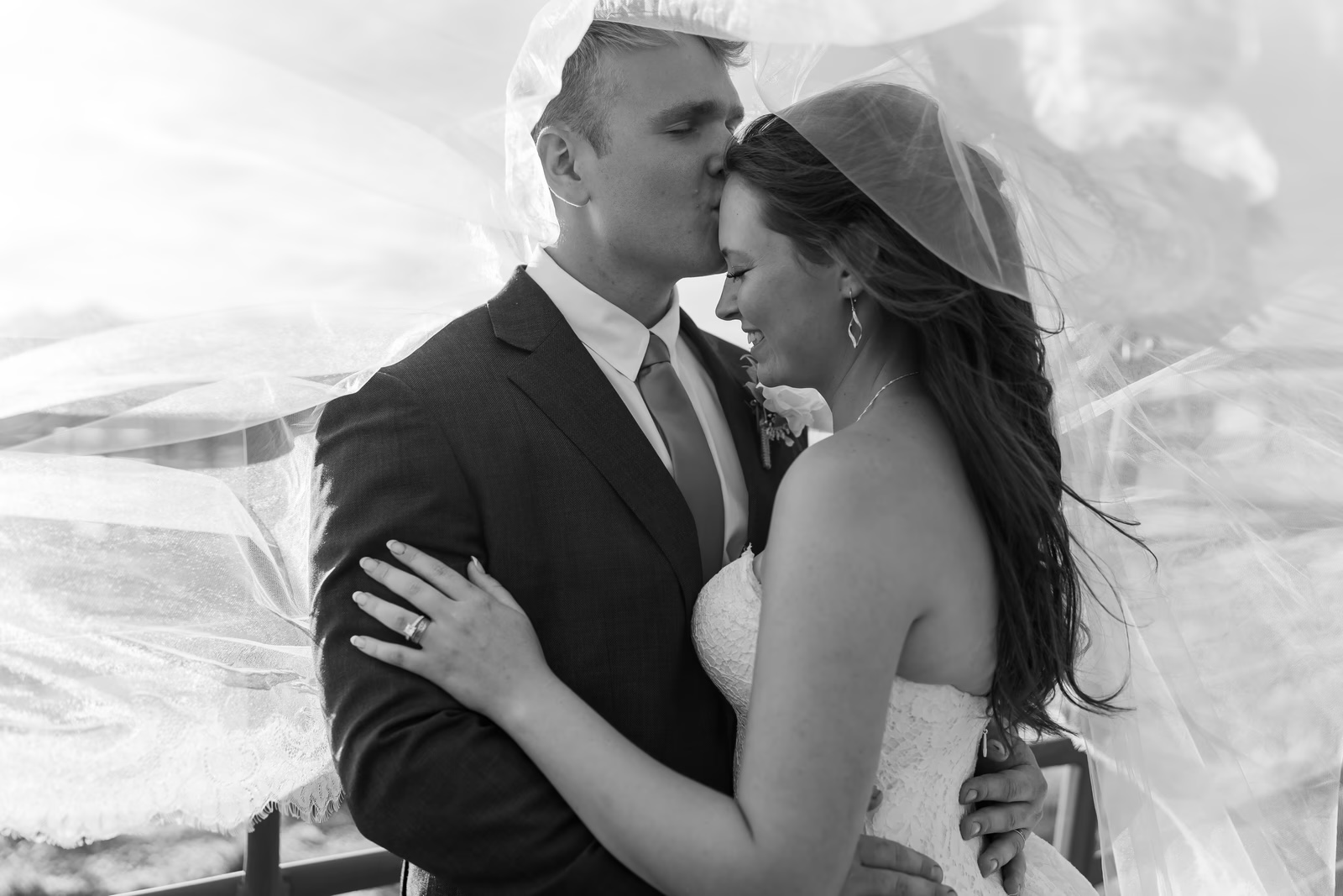 Black-and-white portrait of a groom kissing the bride’s forehead beneath her veil