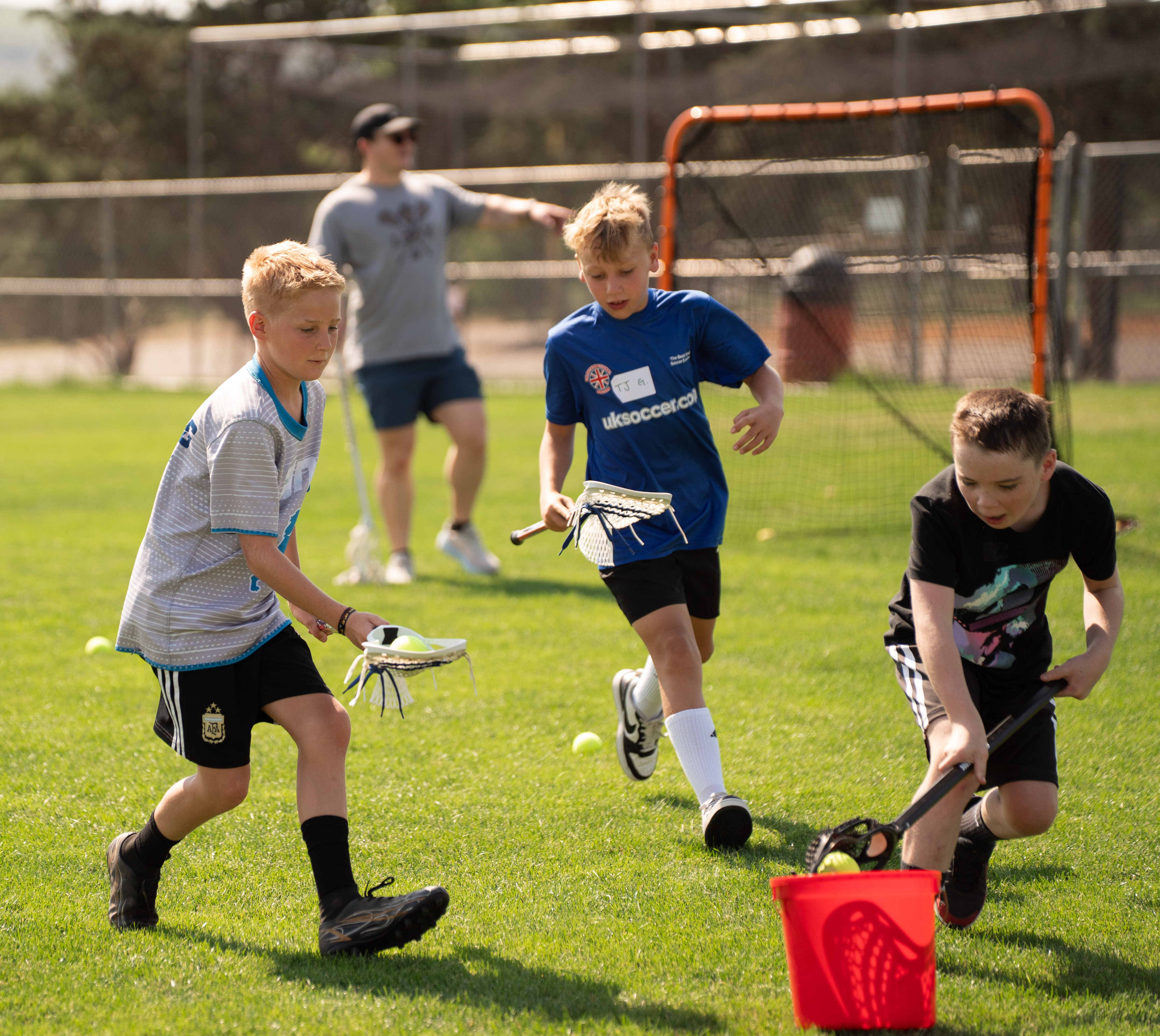 three children with lacrosse sticks place balls in an orange bucket on a field with a lacrosse goal and a coach behind them