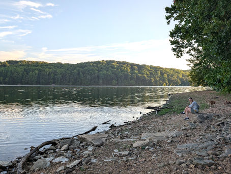 A person sits with a dog on the rocky shore of Loch Raven Reservoir as evening light reflects across the water.