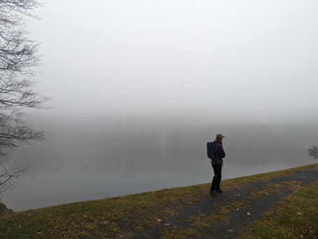 A person in a dark jacket and cap stands alone on a grassy bank at the edge of a still lake, wearing a backpack and looking out toward a far shore that is mostly hidden by heavy gray fog.