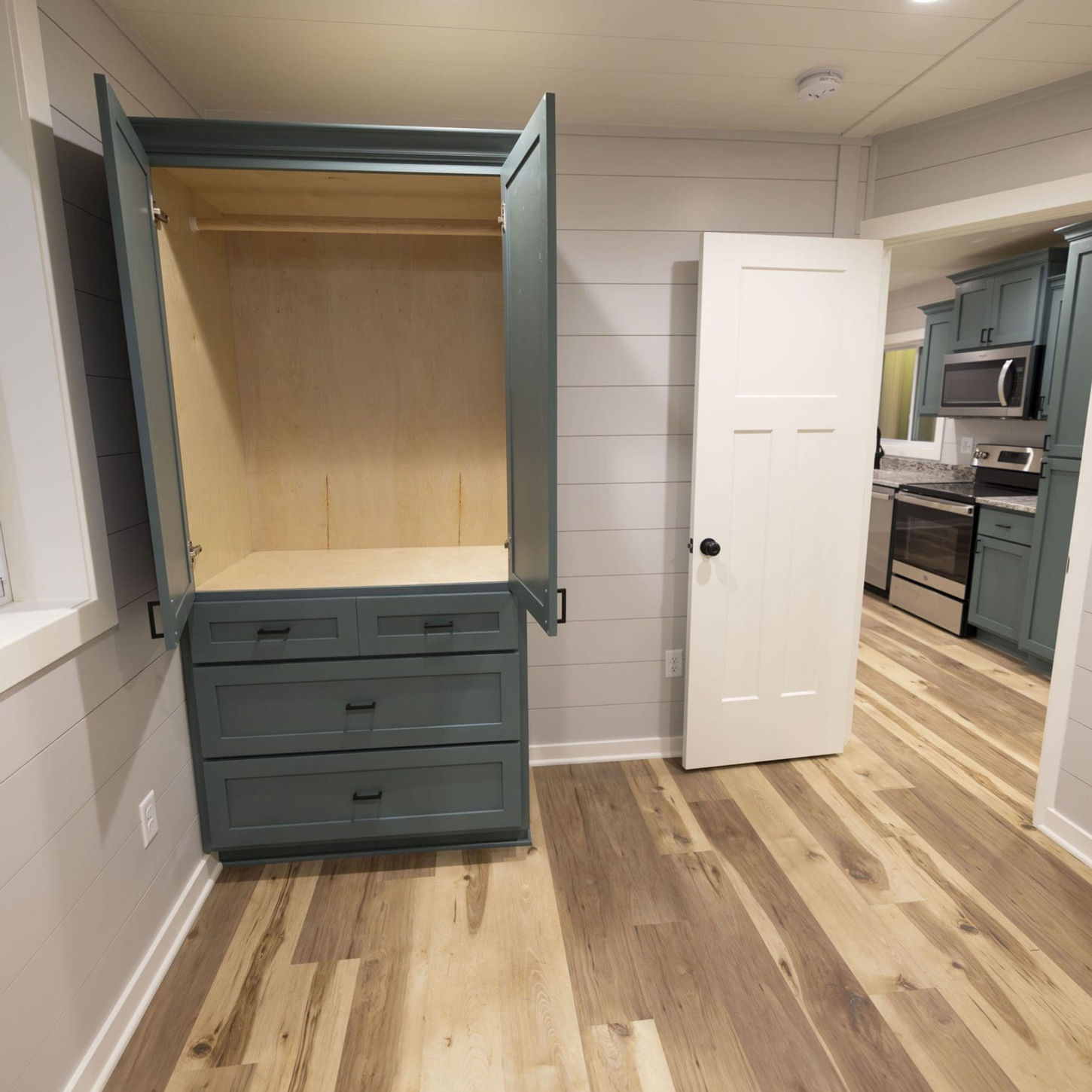 Compact bedroom featuring open built-in wardrobe, leading to tiny home kitchen.