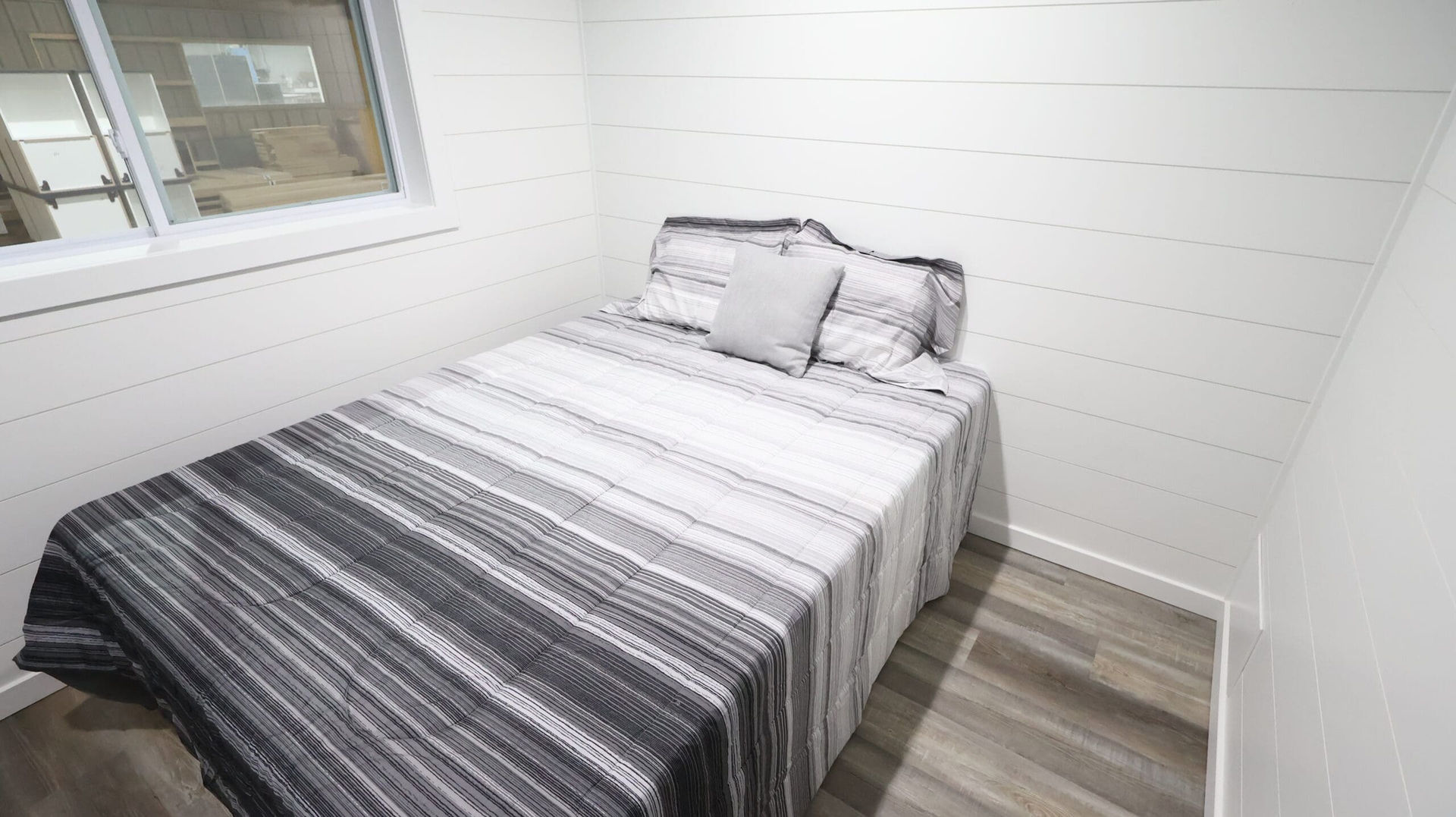 Small bedroom with bed, gray striped bedding, shiplap walls, wood floor.