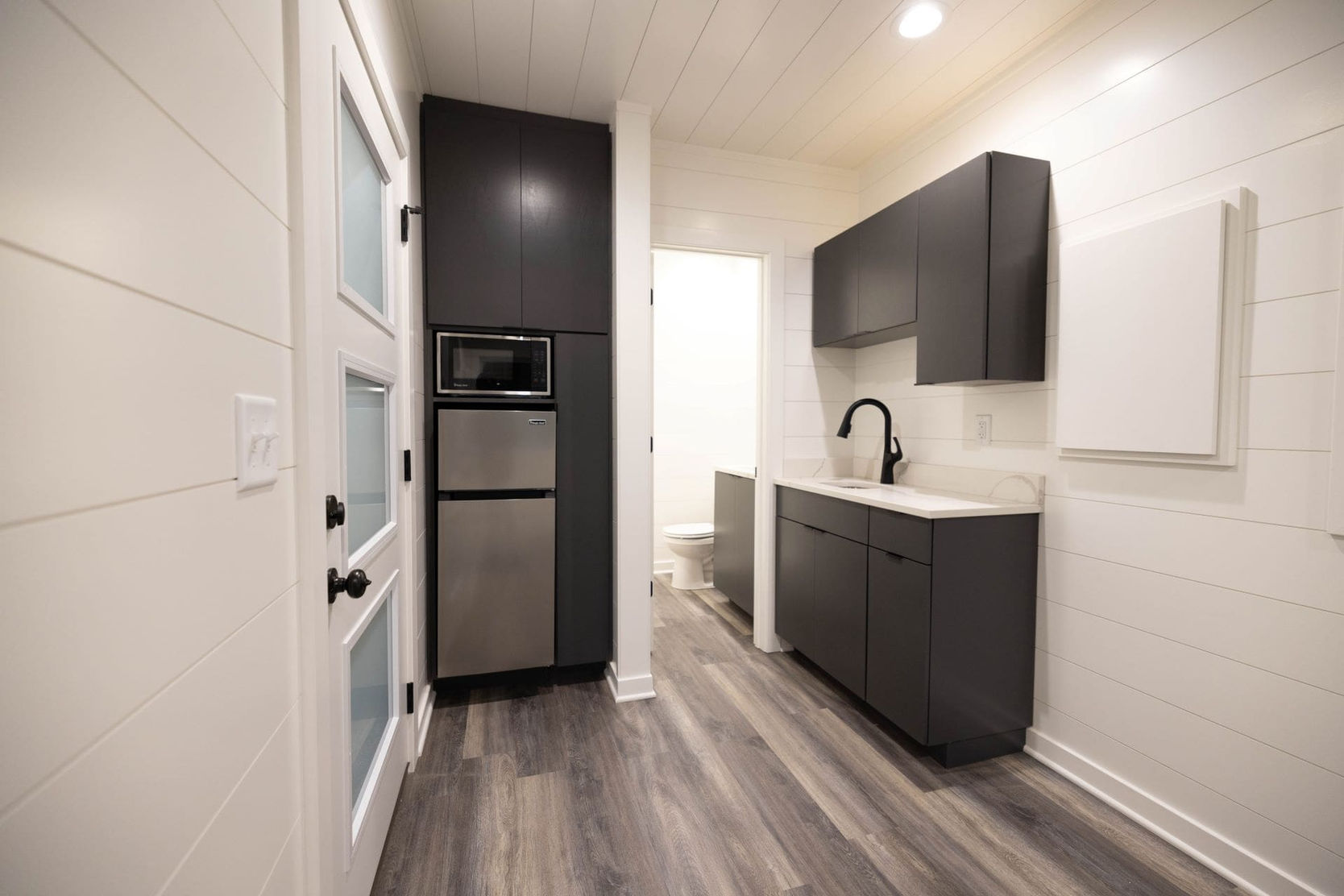 Modern kitchenette with dark cabinets, white counter, sink, microwave, refrigerator.