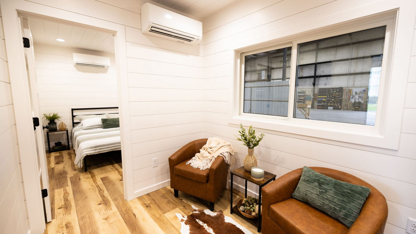 Modern tiny home interior with brown armchairs, wood floor, and bedroom.
