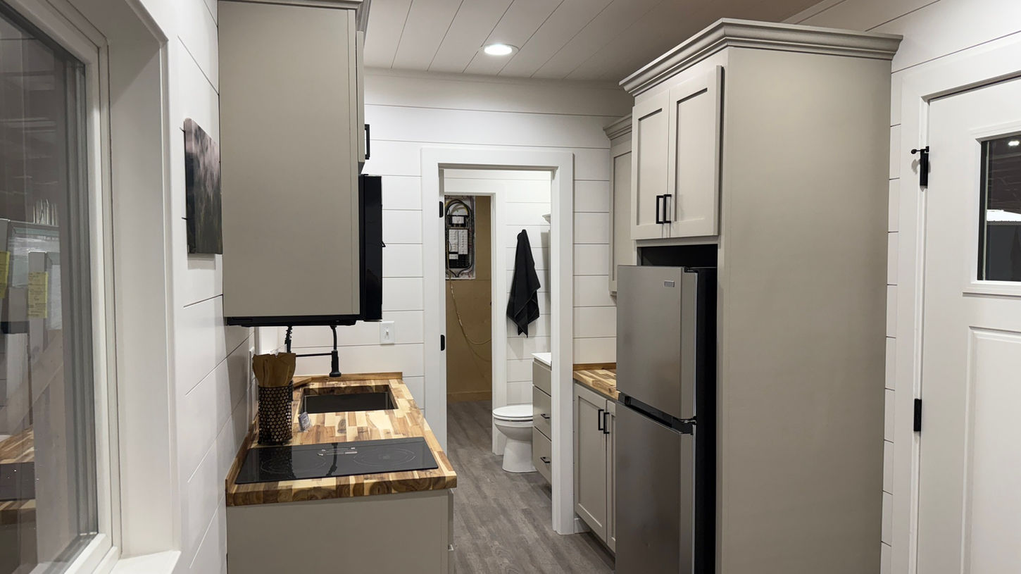 Narrow tiny home kitchen with light gray cabinets, wood counters, and appliances.