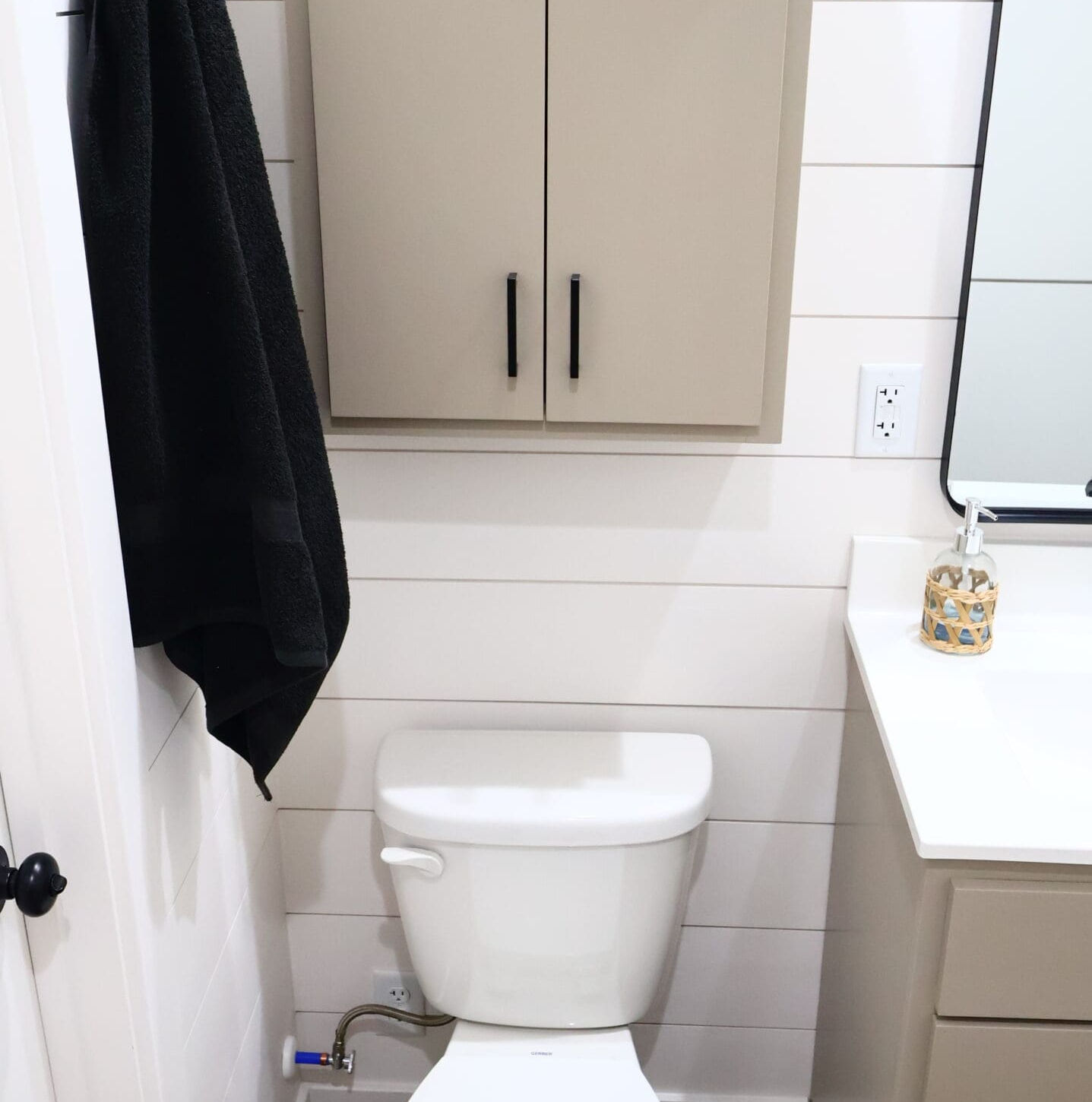 Toilet in small bathroom with shiplap walls, beige cabinet, black towel.