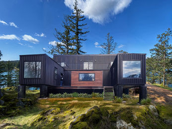 Dark modern house on stilts, rocky shore, trees, blue sky.