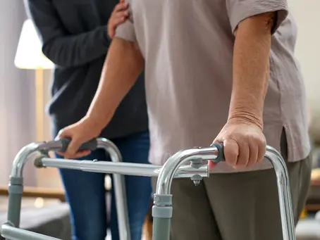 Elderly person using a walker with caregiver support in a home setting.