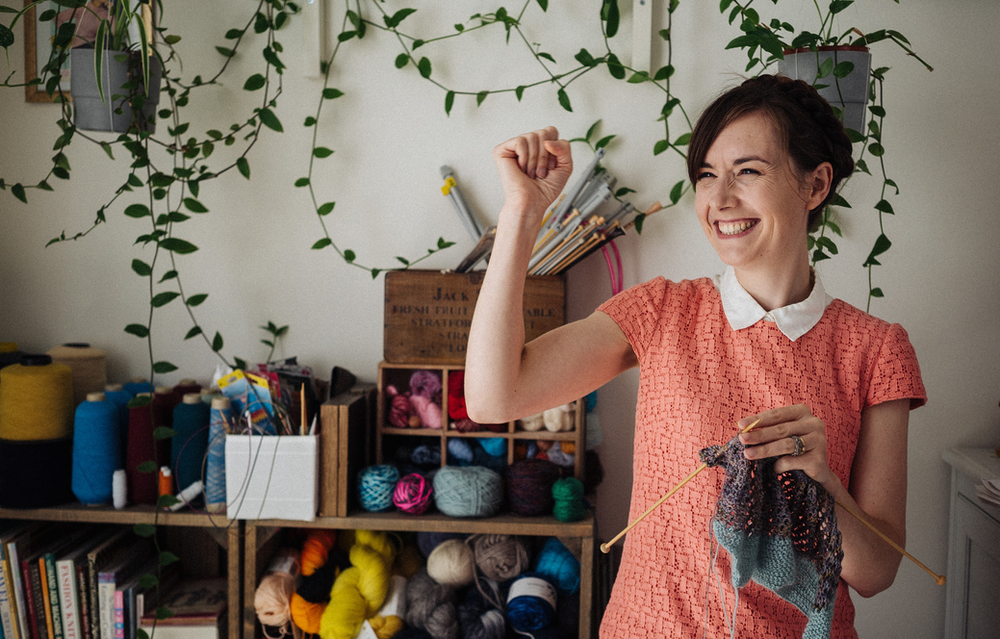 A white woman in her 30's has knitting in her right hand and her left hand up in a fist, celebratory gesture. She wears a peach dress with white collar and has knitting needles and yarn and green vines behind her.