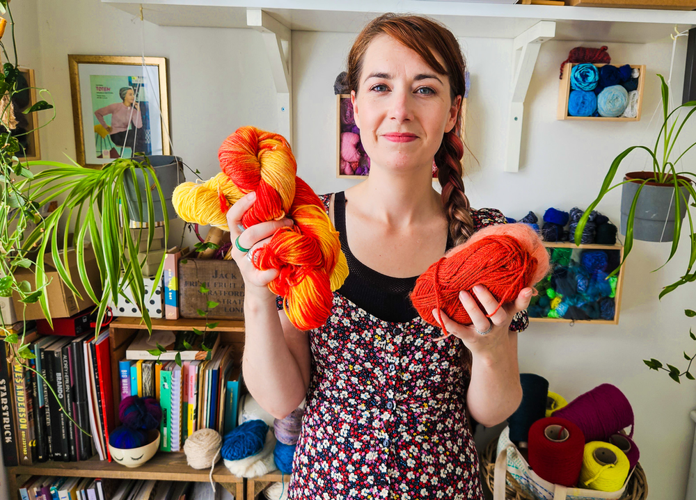 A white female knitting teacher holds orange coloured yarn in each hand