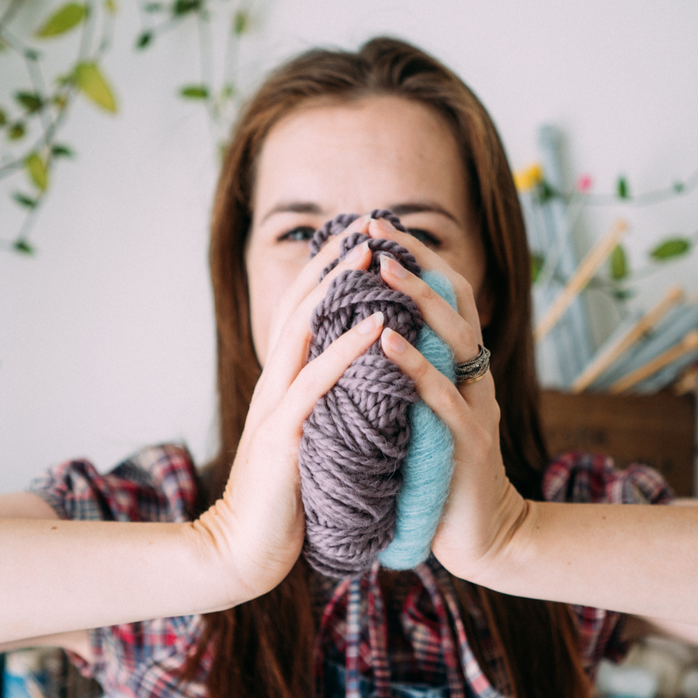 A white woman, Alice, a knitting teacher. Squashes 2 different yarn balls together in front of her face.