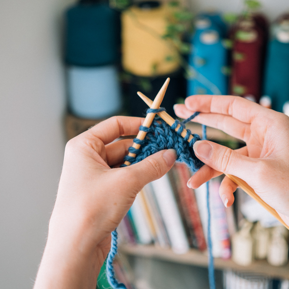 a white woman's hands hold wooden knitting needles with blue knitting on it. She is mid row.