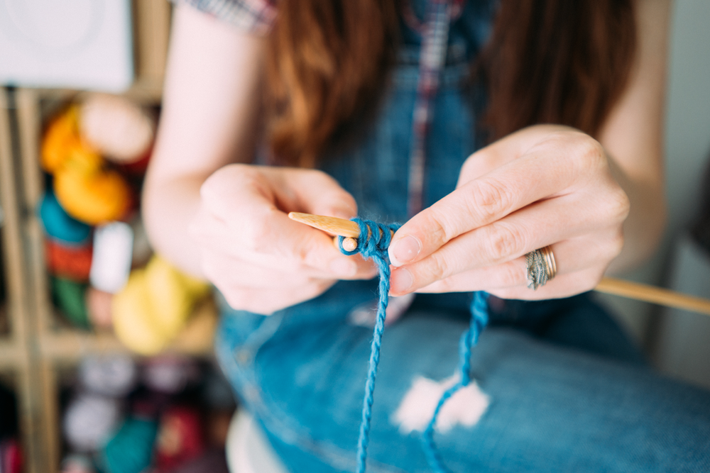 A white woman's hands hold wooden knitting needles with blue yarn on them