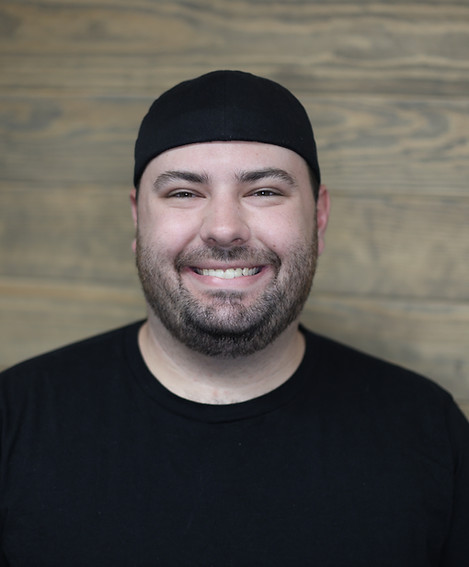 Drake's headshot. The creator of this site stands in front of a wooden background, smiling at the camera in a black shirt, with a black ballcap on his head. 