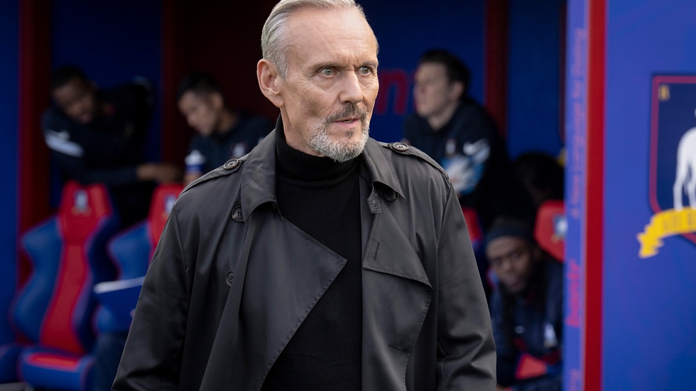 Man in black coat (Rupert Mannion) stands in front of a sports bench. Background includes blurred people in blue and red seats, suggesting a serious mood.