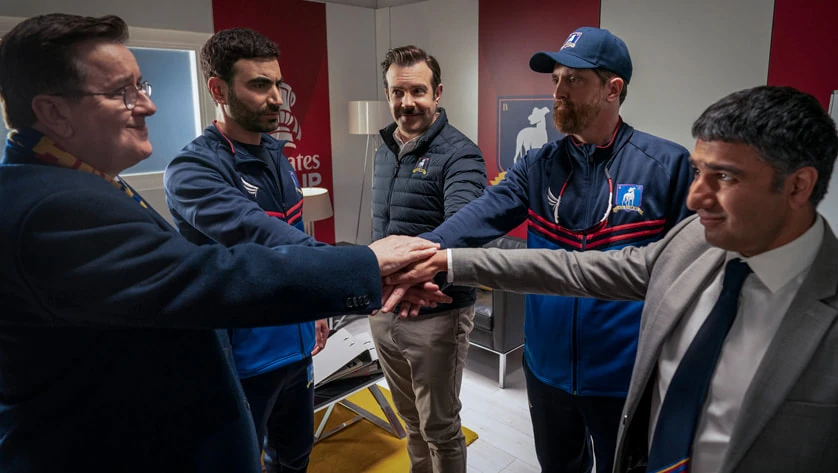 Five men (Ted Lasso's Diamond Dogs - Ted, Beard, Nathan, Higgins, and Roy, Trent is missing) stand in a room, hands stacked together, showing unity. They wear casual and sports attire. The room has red and white walls.