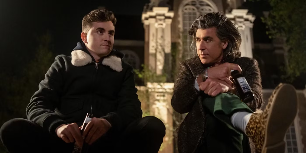 Two men (Colin and Trent) sit outdoors at night, holding bottles. Colin wears a black jacket with white trim, Trent in a patterned coat and leopard shoes. Moody setting.