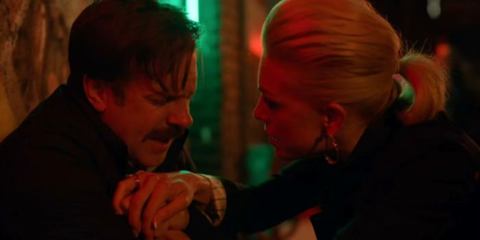 Rebecca sits with Ted to support him during a panic attack. They are engaged in a tense conversation in a dimly lit setting with red and green lights. Rebecca appears supportive, holding Ted's hand.
