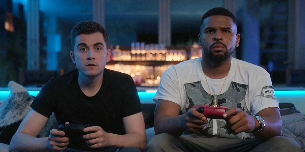 Two men (Colin and Isaac) focused on playing video games with controllers in a dimly lit room. Blue lighting and shelves of bottles in the background.