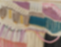A close up of a macrame wall art created using knitted sections , crochet, fringing and tassles in shades of cream, pink and teal blue