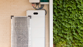 A wall-mounted home battery energy storage system installed on a beige brick exterior wall, with white modular units stacked vertically and connected by black cables to an inverter unit above. A dusty filter or mesh screen is visible on the left side of the unit. Lush green ivy covers the adjacent wall on the right side of the image