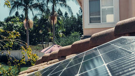 A close-up view of solar panels installed on a tiled house roof. In the background, there are palm trees, a garden, and part of a peach-coloured house with a white-framed window. The sunlight is reflecting off the smooth surface of the solar panels.