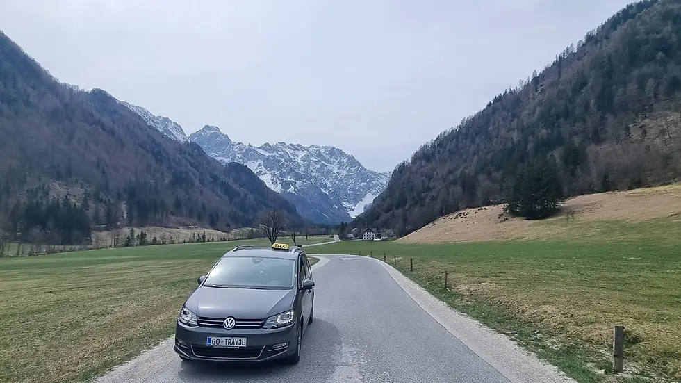 Samochód z napisem "TAXI" na drodze w górskiej dolinie w Słowenii, ośnieżone szczyty w tle. Tablica rejestracyjna GO TRAV3L.