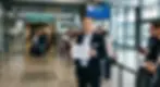 A man in a suit waits in the arrivals hall with a sign reading "Welcome Anna Kowalska." A crowd of passengers and a flight information board are in the background. Gdańsk.