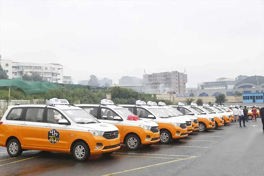 Szereg żółto-białych taksówek na etloskim parkingu, obok kilku ludzi. W tle budynki, niebo pochmurne. Na taksówkach napis "TAXI".