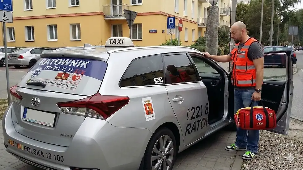 A man in a life jacket stands next to a silver car with taxi and lifeguard markings. A yellow building stands in the background. He is holding a red backpack.