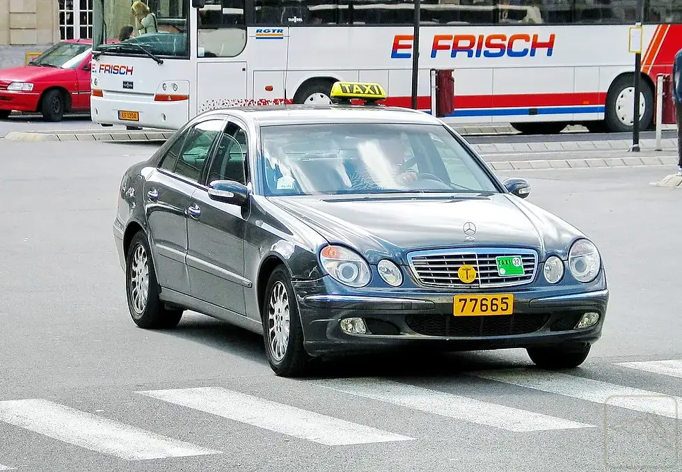 Ein graues Mercedes-Taxi mit der Nummer 77665 steht am Kreuzungspunkt, im Hintergrund ein E FRISCH-Bus und ein rotes Auto.
