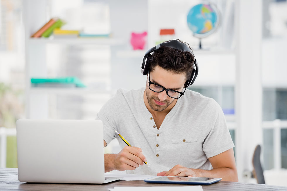 Man wearing headphones and glasses writes at a desk beside a laptop and tablet in a bright study room with a globe behind him.