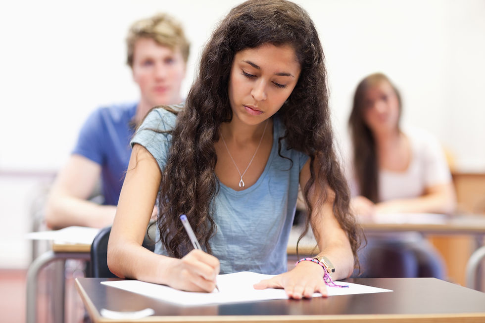Focused student with long dark hair writes on paper in a classroom, with two blurred classmates in the background.