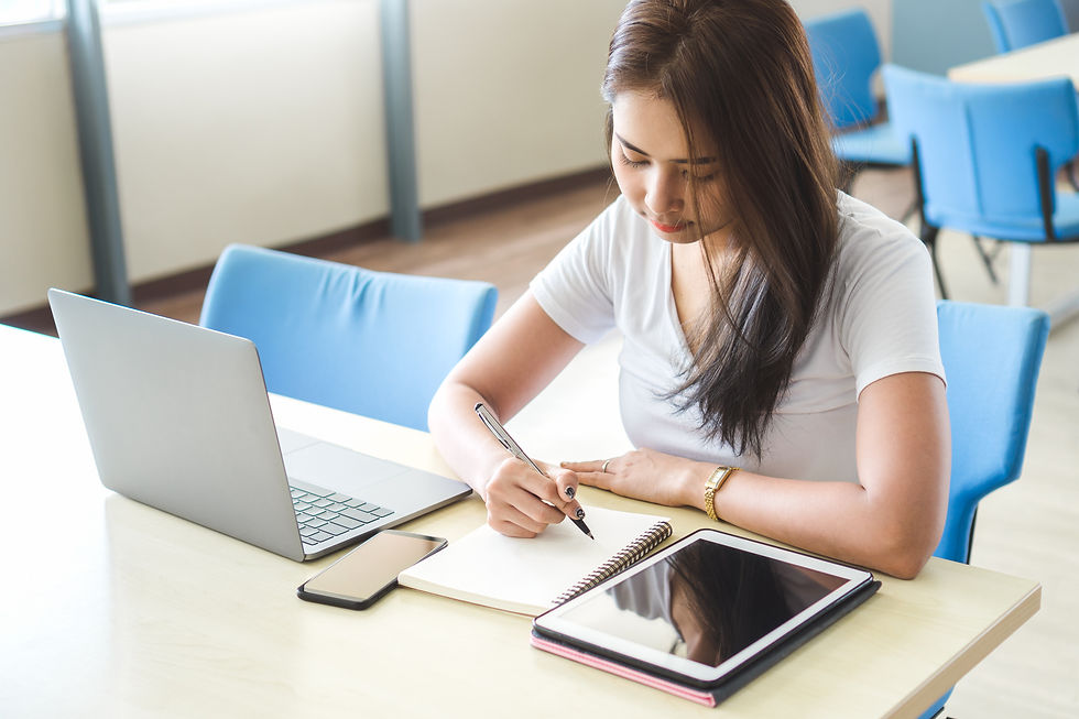 Young woman writes in a notebook beside an open laptop, phone, and tablet in a bright room with blue chairs.