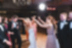 Bride dancing joyfully at a wedding, surrounded by guests in formal attire. A lively reception atmosphere with bright lighting.