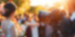 Bride with floral hairpiece stands before a camera at sunset. Blurred crowd and warm colors create a dreamy, romantic atmosphere.