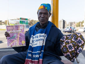 Big Issue vendor posing at the traffic lights with the magazine in one hand and his cap in another. He is happy and jovial.