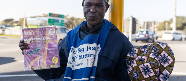 Big Issue vendor posing at the traffic lights with the magazine in one hand and his cap in another. He is happy and jovial.