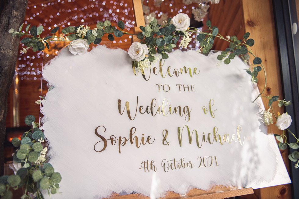 A white and gold sign welcoming guests to the wedding of Sophie and Michael at a wedding at Canada Lodge and Lake, Cardiff, photographed by Swansea Wedding Photographer, Katy Tainton Photography