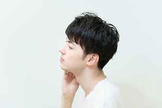 白い背景で横向きに座り、顎に手を添えている短髪の男性