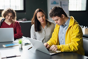 Young male with Down syndrome uses a laptop. A woman is sitting next to him and talking to him as he types. 