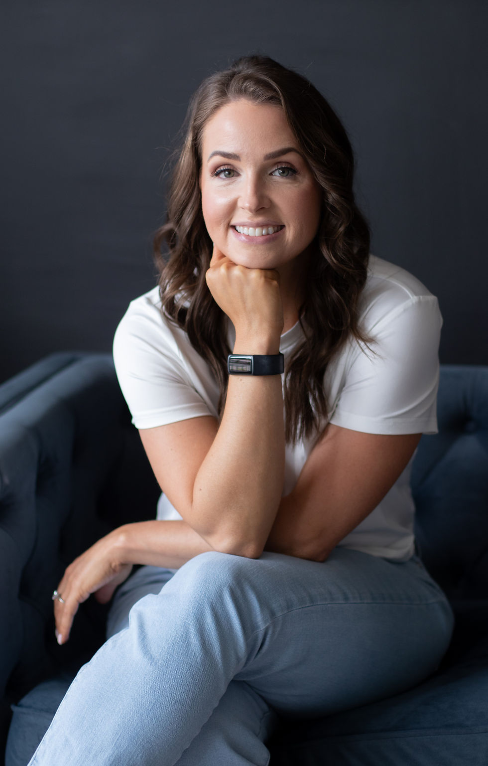 Smiling woman with wavy hair sits on a blue couch, wearing a white shirt and a smartwatch. Black background, relaxed and cheerful mood.