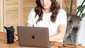 A woman in a white shirt works on a laptop at a wooden table, holding a black tumbler. Phone and glasses are beside her; tropical plants in the background.