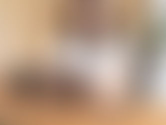 A woman in a white shirt works on a laptop at a wooden table, holding a black tumbler. Phone and glasses are beside her; tropical plants in the background.