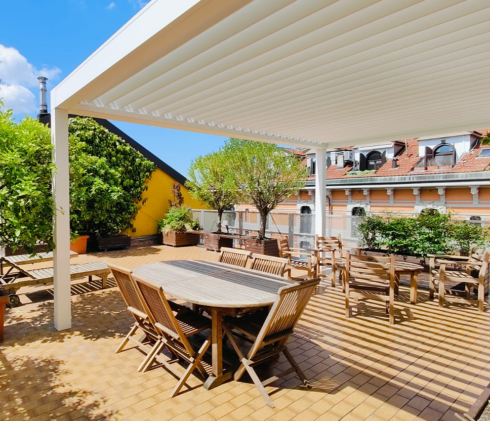 Pergola bioclimatica addossata a parete. La struttura è totalmente in alluminio, color bianco. Perimetralmente la pergola non ha chiusure, ma c'è la predisposizione per le tende verticali e le vetrate scorrevoli. la apergola bioclimatica è installata su una grande terrazza di un palazzo di Milano. Sotto alla pergola c'è il classico tavolo con sedie e divano in legno per esterno. Il terrazzo ha grandi vasi che contengono piante di limoni e gelsomini