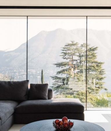 Serramento scorrevole minimale in alluminio. E' installato in un salotta. Il serramento si affaccia su un balcone con parapetto in vetro. Il panorama ha come sfondo una montagna