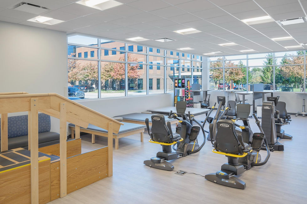 Rehabilitation gym at the PACE KC Adult Wellness Center by Swope Health featuring therapy equipment, recumbent exercise bikes, treatment tables, and mobility training stairs overlooking large windows.