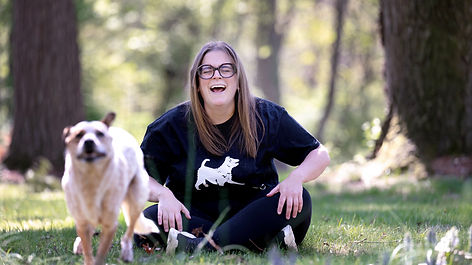 a brunette woman sits on the ground laughing as a blurry dog races towards the camera